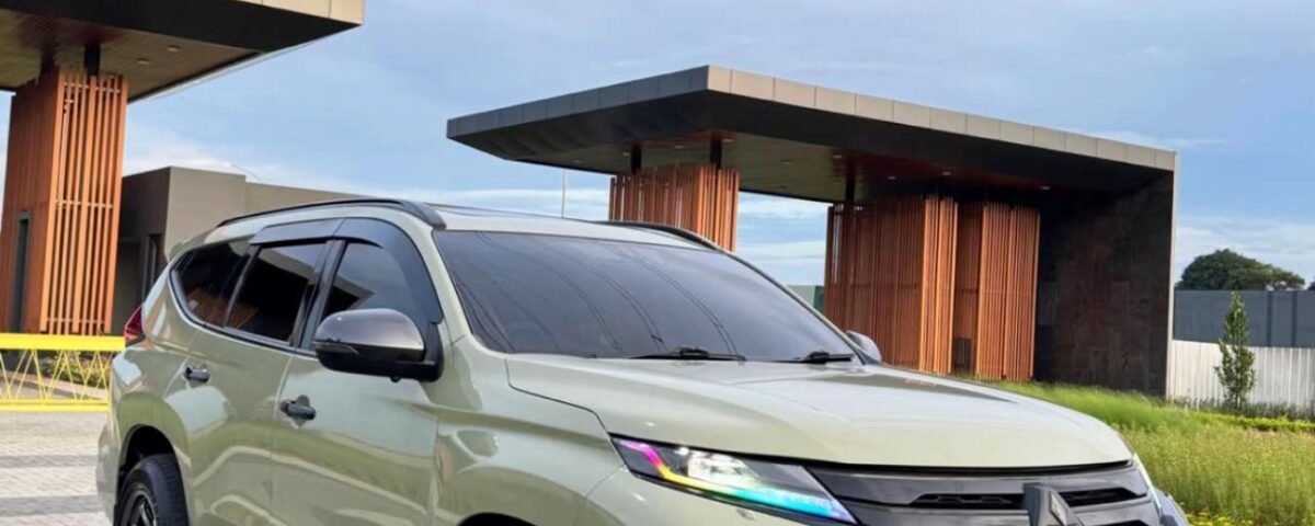 Light green Mitsubishi SUV parked on a brick driveway near modern orange-columned buildings under a blue sky with overhead power lines.