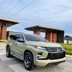 Light green Mitsubishi SUV parked on a brick driveway near modern orange-columned buildings under a blue sky with overhead power lines.