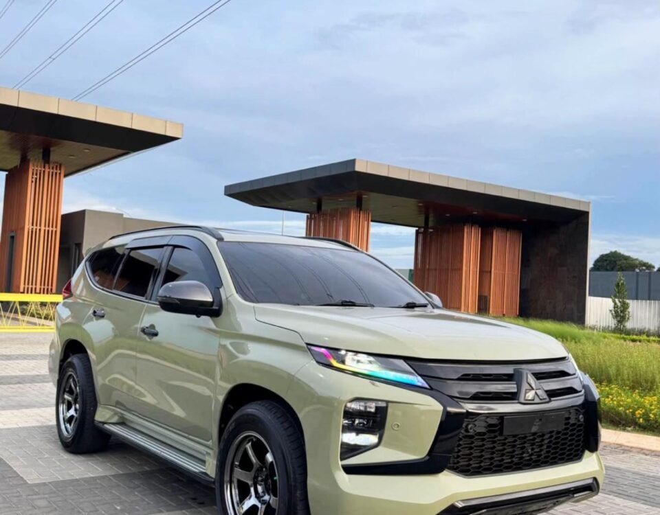 Light green Mitsubishi SUV parked on a brick driveway near modern orange-columned buildings under a blue sky with overhead power lines.