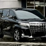 Black luxury minivan parked on a wet, reflective pavement outside a modern building with large glass windows.