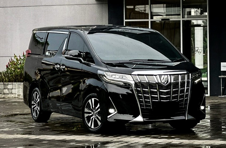 Black luxury minivan parked on a wet, reflective pavement outside a modern building with large glass windows.