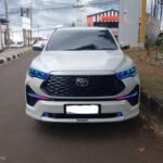 snaptik_7593942426075680007_1_v2 - Dyantransport Front view of a white Toyota SUV parked on a street, blue LED headlights glowing, urban backdrop with power lines.