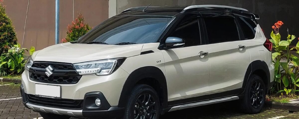 White Suzuki SUV parked in a shaded area beside a brown wall, with plants on the left.