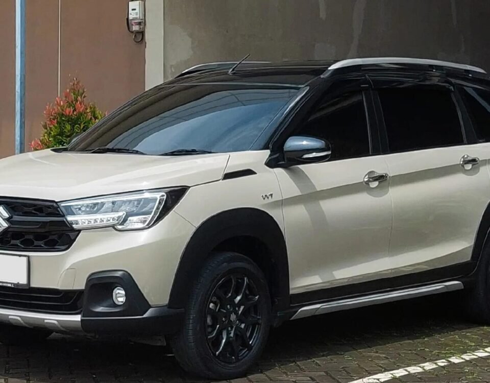 White Suzuki SUV parked in a shaded area beside a brown wall, with plants on the left.