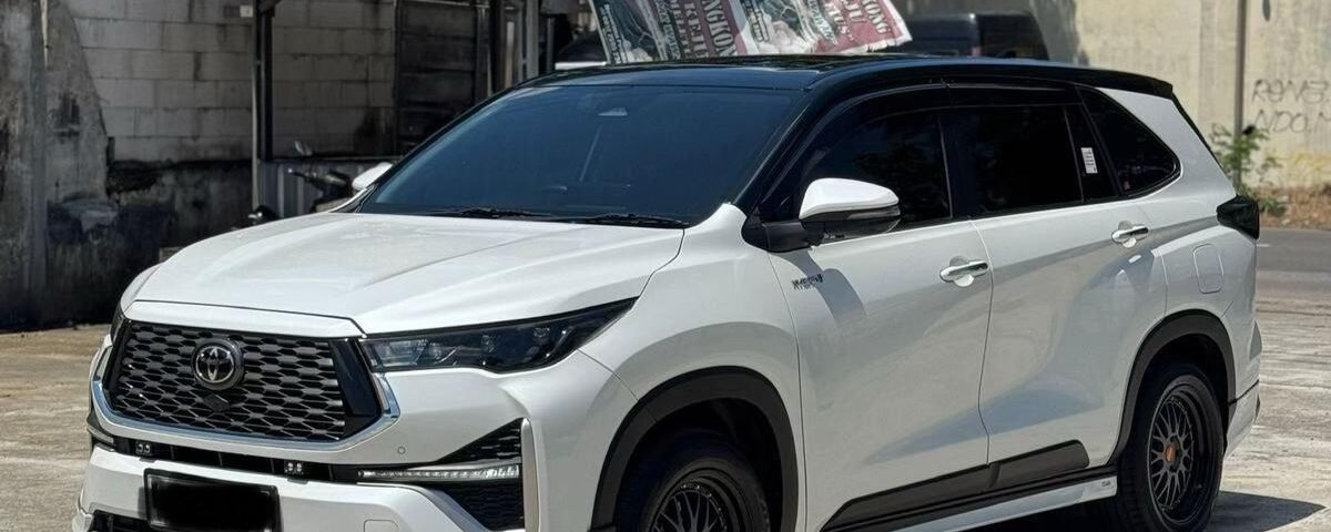 White Toyota SUV parked outdoors on a sunny day, with a large banner draped over its roof. Trees and an industrial building are in the background.