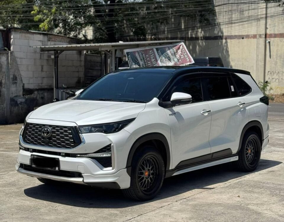 White Toyota SUV parked outdoors on a sunny day, with a large banner draped over its roof. Trees and an industrial building are in the background.