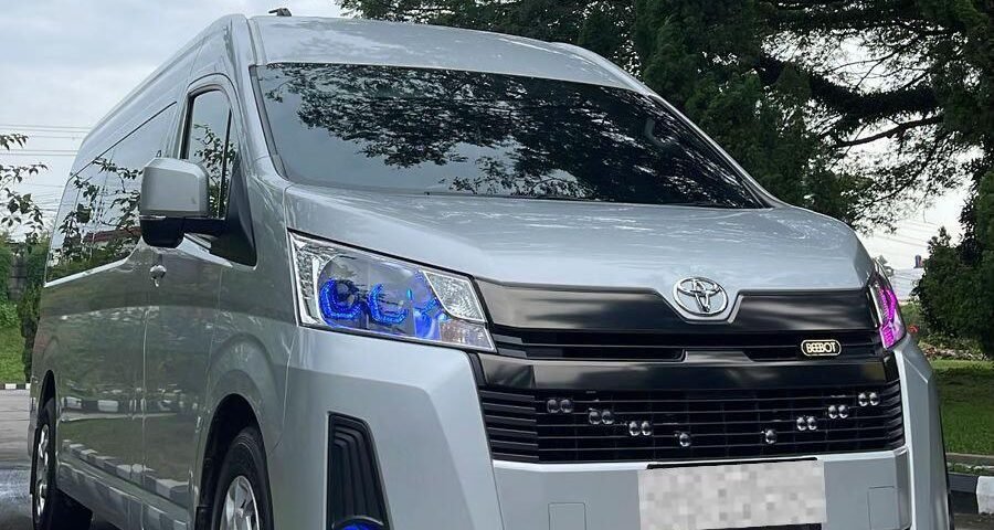 Front three-quarter view of a silver Toyota van parked on a paved road beneath trees with blue-tinted headlights visible.