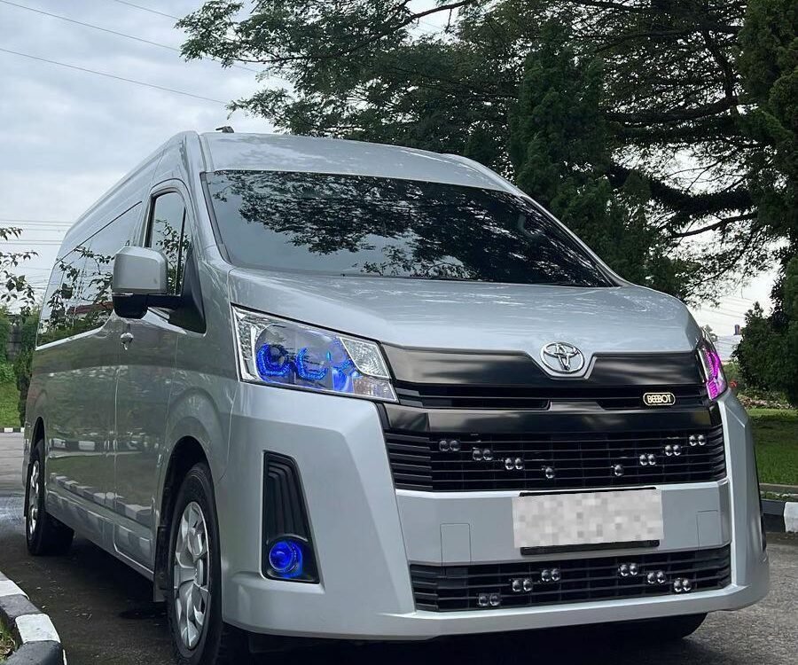 Front three-quarter view of a silver Toyota van parked on a paved road beneath trees with blue-tinted headlights visible.