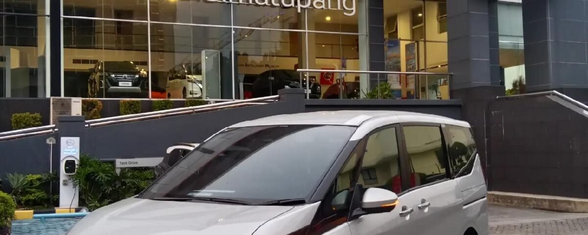 SaveTikTokto_7491337582182141192_2 - Dyantransport Silver Nissan minivan parked in front of a Nissan dealership with a glass showroom and large red Nissan sign above the entrance.
