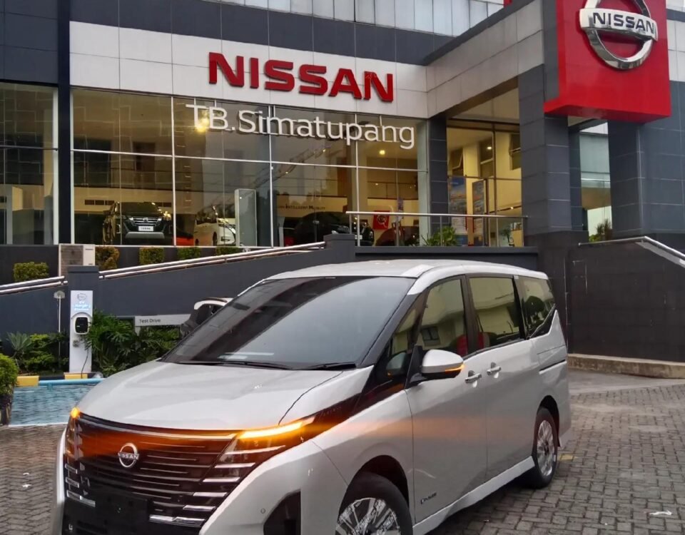 Silver Nissan minivan parked in front of a Nissan dealership with a glass showroom and large red Nissan sign above the entrance.