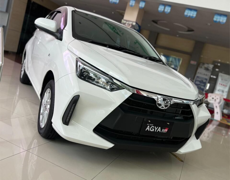 White Toyota Agya GR Sport on a showroom floor, angled front view with glossy tiles and showroom signage in the background