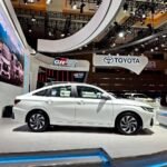 SaveTikTokto_7603938007632170258_3 - Dyantransport White sedan on a pedestal at an auto show, with a prominent Toyota booth and futuristic curved architecture overhead