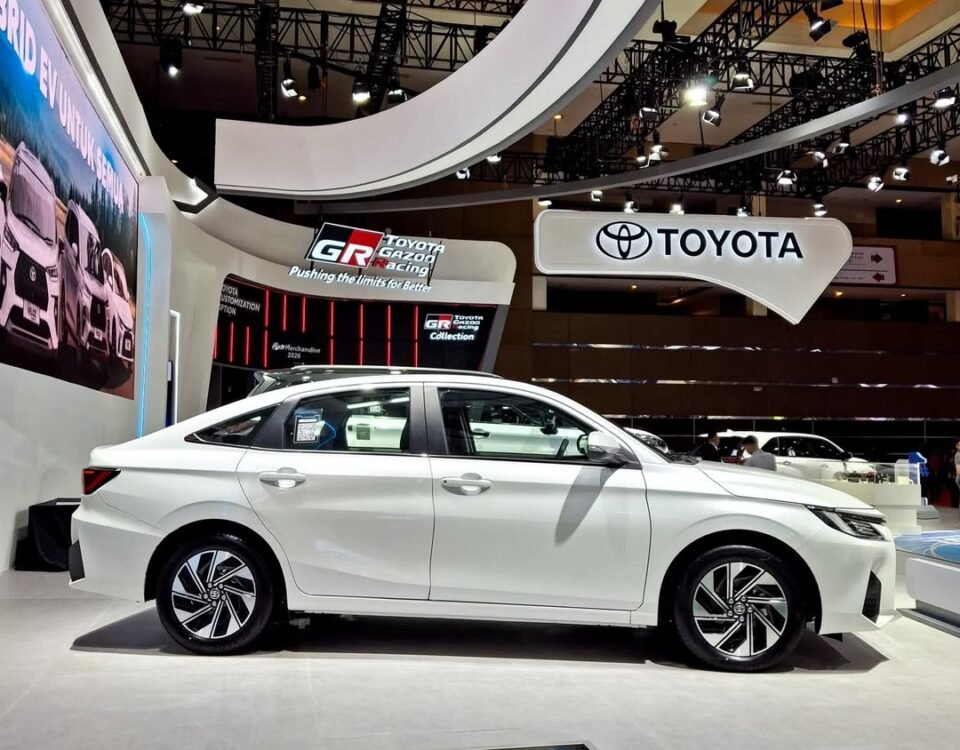 White sedan on a pedestal at an auto show, with a prominent Toyota booth and futuristic curved architecture overhead