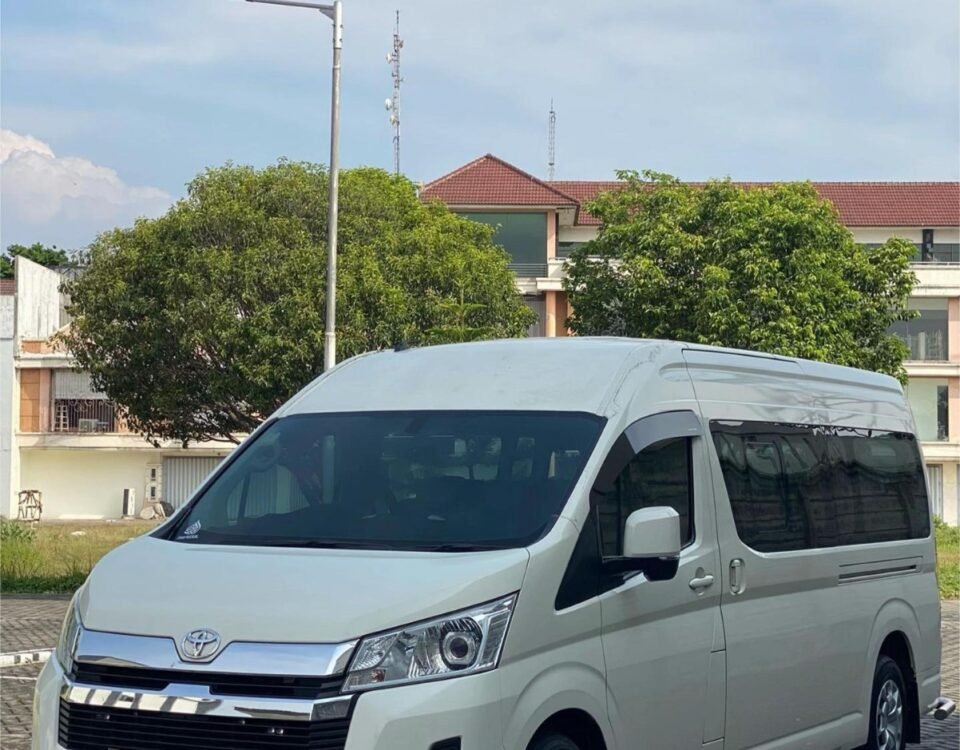 Front three-quarter view of a white Toyota van parked on a brick-paved lot with trees and buildings in the background.