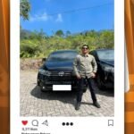 Man in a beige shirt and sunglasses posing in front of two black SUVs in a sunny outdoor lot with greenery behind him from a social post image.
