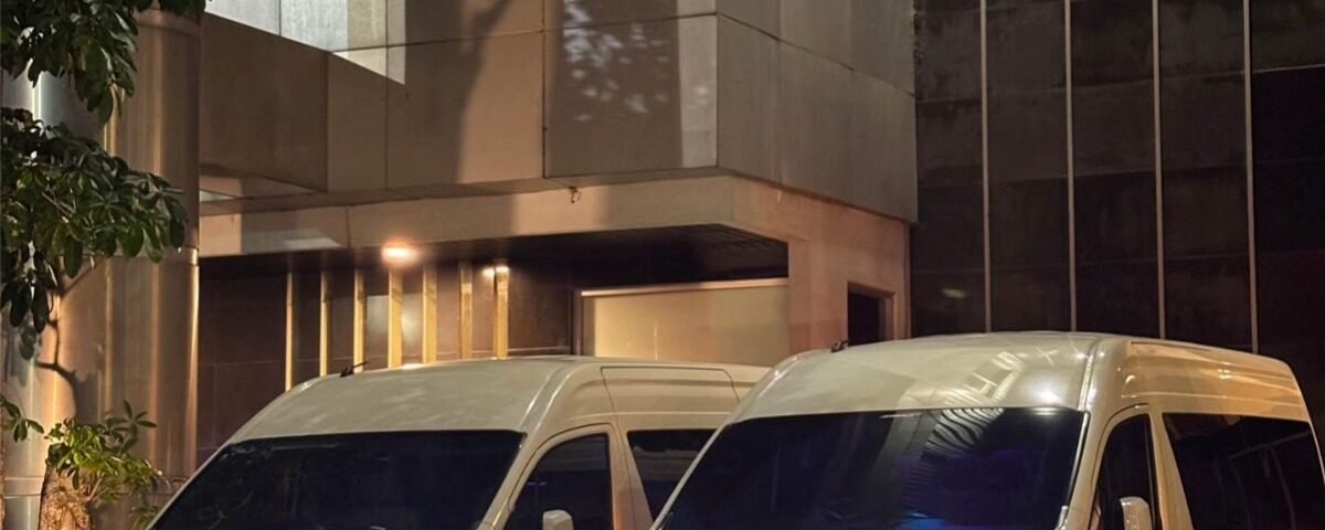 WhatsApp Image 2026-08-22 at 153347 - Dyantransport Two white vans parked side by side outside a modern glass-and-steel building at night, with headlights on.
