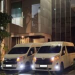 WhatsApp Image 2026-08-22 at 153347 - Dyantransport Two white vans parked side by side outside a modern glass-and-steel building at night, with headlights on.