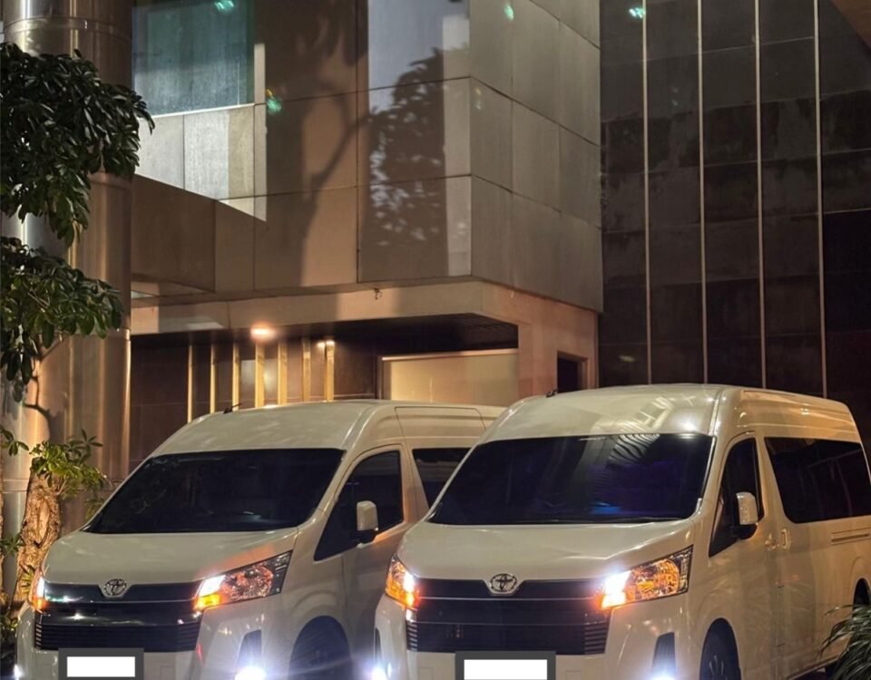 WhatsApp Image 2026-08-22 at 153347 - Dyantransport Two white vans parked side by side outside a modern glass-and-steel building at night, with headlights on.