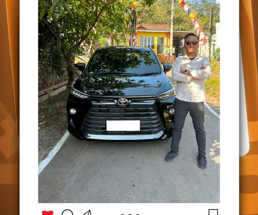 Man standing with arms crossed beside a black Toyota SUV in a parking area with festive bunting