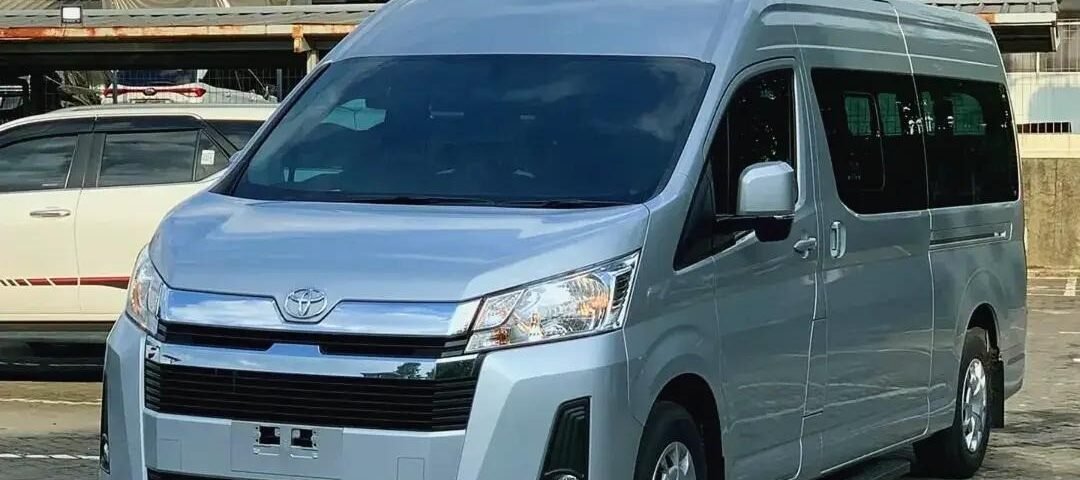 Front three-quarter view of a silver Toyota van parked in a paved urban parking lot with buildings in the background.