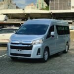 premio 12121212 - Dyantransport Front three-quarter view of a silver Toyota van parked in a paved urban parking lot with buildings in the background.
