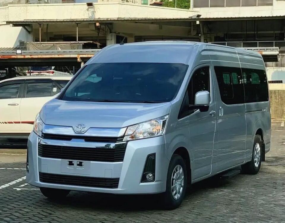 Front three-quarter view of a silver Toyota van parked in a paved urban parking lot with buildings in the background.