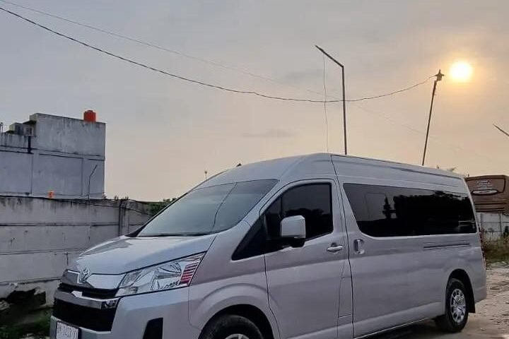 snaptik-app-7442597708428348727-slide-3 - Dyantransport White modern van parked on a dirt lot beside a low wall, with a sunset sky and streetlight in the background.