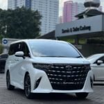 snaptik-app-7512311422894755128-slide-2 - Dyantransport White Toyota Alphard minivan in a parking lot with a dealership sign in the background and tall buildings behind.