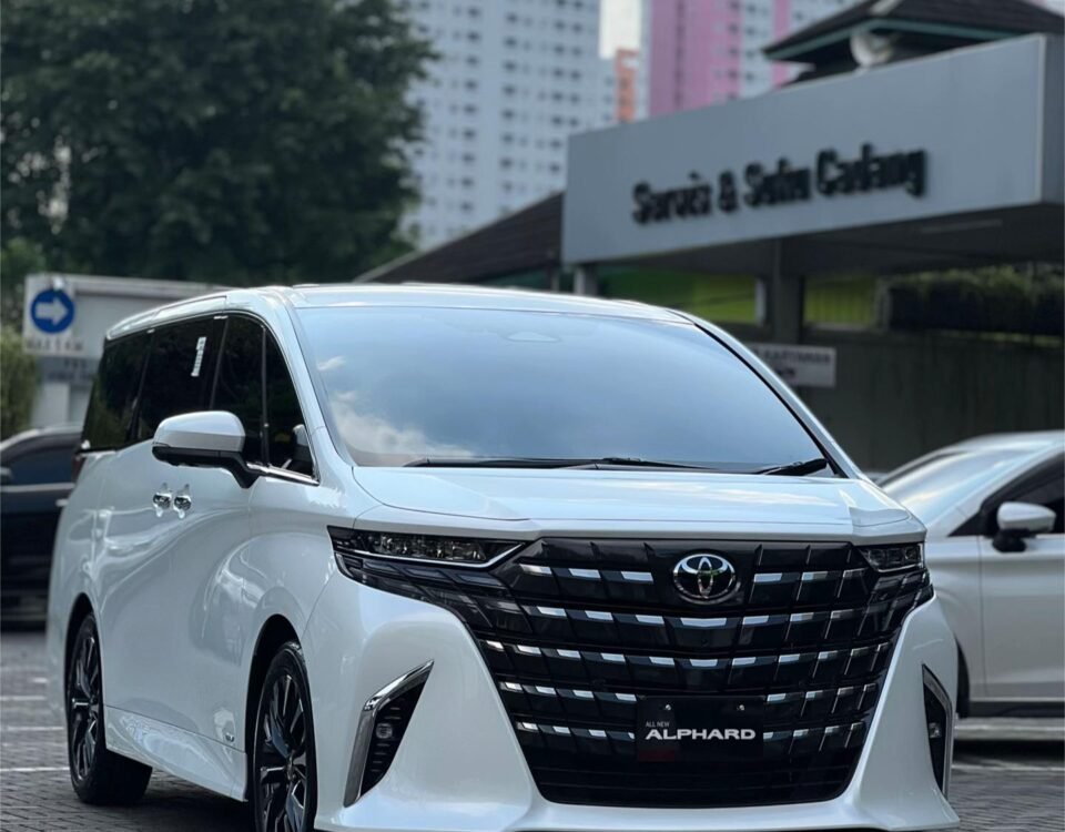 White Toyota Alphard minivan in a parking lot with a dealership sign in the background and tall buildings behind.