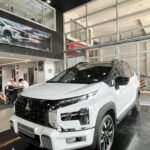 White Mitsubishi Xpander Cross SUV on display in a dealership showroom with glass-walled offices behind it.