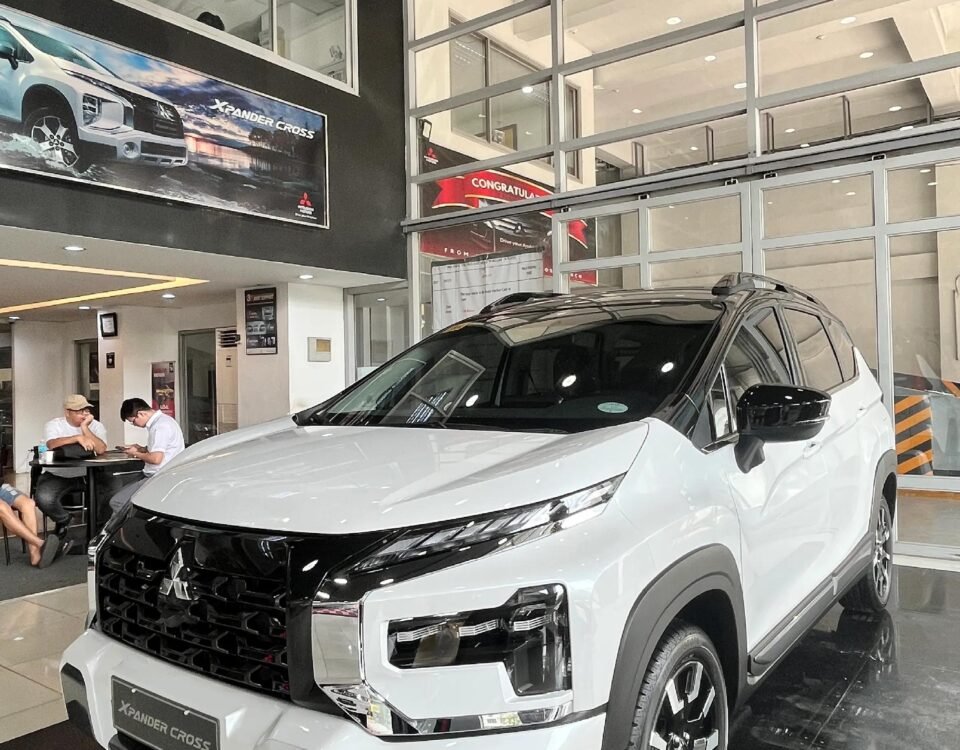 White Mitsubishi Xpander Cross SUV on display in a dealership showroom with glass-walled offices behind it.