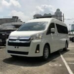 snaptik-app-7577675428547136789-slide-1 - Dyantransport White Toyota van parked in a lot with other vehicles and urban buildings in the background.