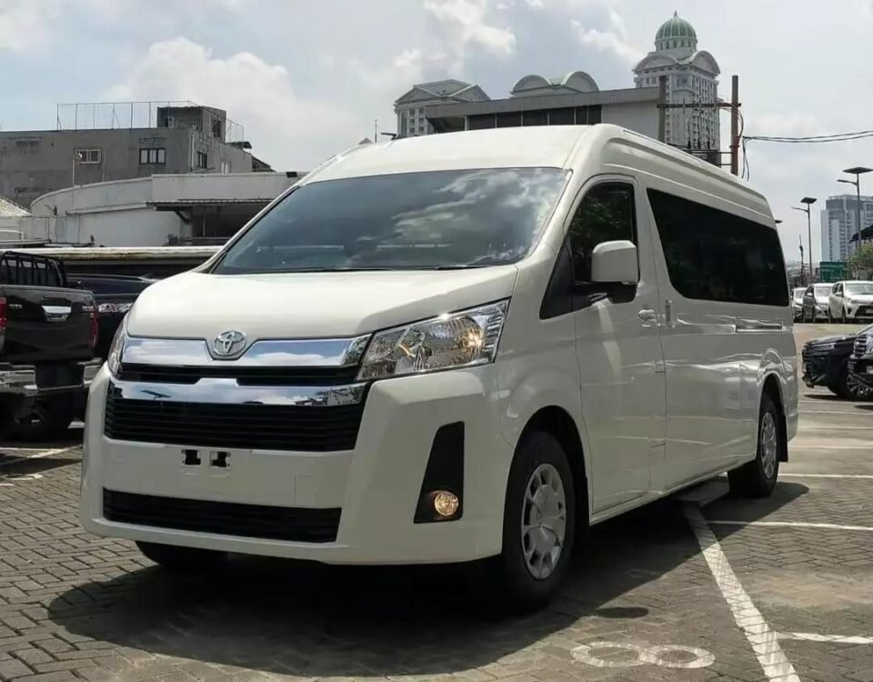 snaptik-app-7577675428547136789-slide-1 - Dyantransport White Toyota van parked in a lot with other vehicles and urban buildings in the background.