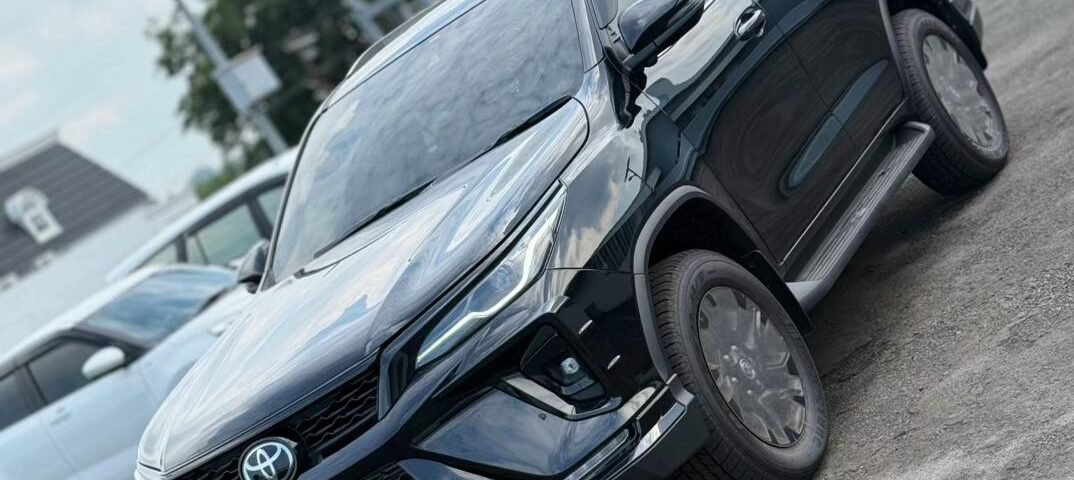 snaptik-app-7590302262082178312-slide-3 - Dyantransport Front-side view of a dark Toyota SUV parked on a lot, angled for a dynamic shot, with the grille and emblem visible.