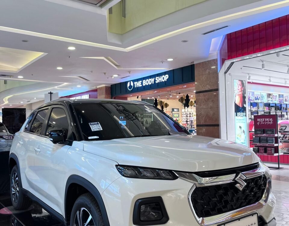 White Suzuki Grand Vitara SUV on a showroom platform inside a shopping mall with The Body Shop storefront in the background.] ,