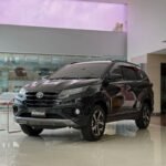Front three-quarter view of a black Toyota SUV on a bright showroom floor, with the 'New Rush GR' badge on the grille.