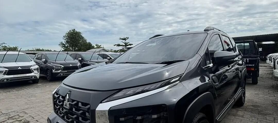 Front three-quarter view of a metallic gray Mitsubishi SUV in a dealership lot with other cars in the background and a cloudy sky above.