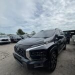 Front three-quarter view of a metallic gray Mitsubishi SUV in a dealership lot with other cars in the background and a cloudy sky above.