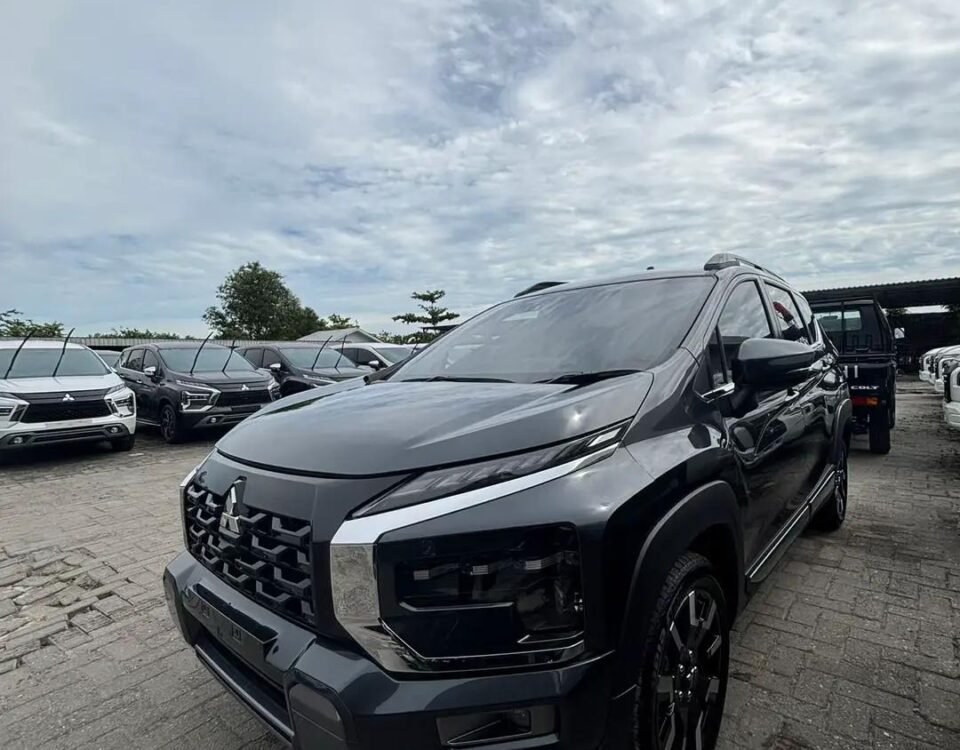 snaptik-app-7609718176263277845-slide-19 - Dyantransport Front three-quarter view of a metallic gray Mitsubishi SUV in a dealership lot with other cars in the background and a cloudy sky above.
