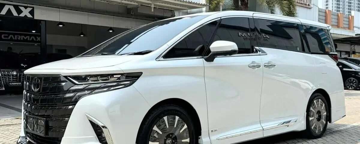 snaptik-app-7647346328464411924-slide-7 - Dyantransport Front three-quarter view of a white luxury minivan with a black grille, parked on a brick driveway in front of a car dealership. Palm trees shade the scene.