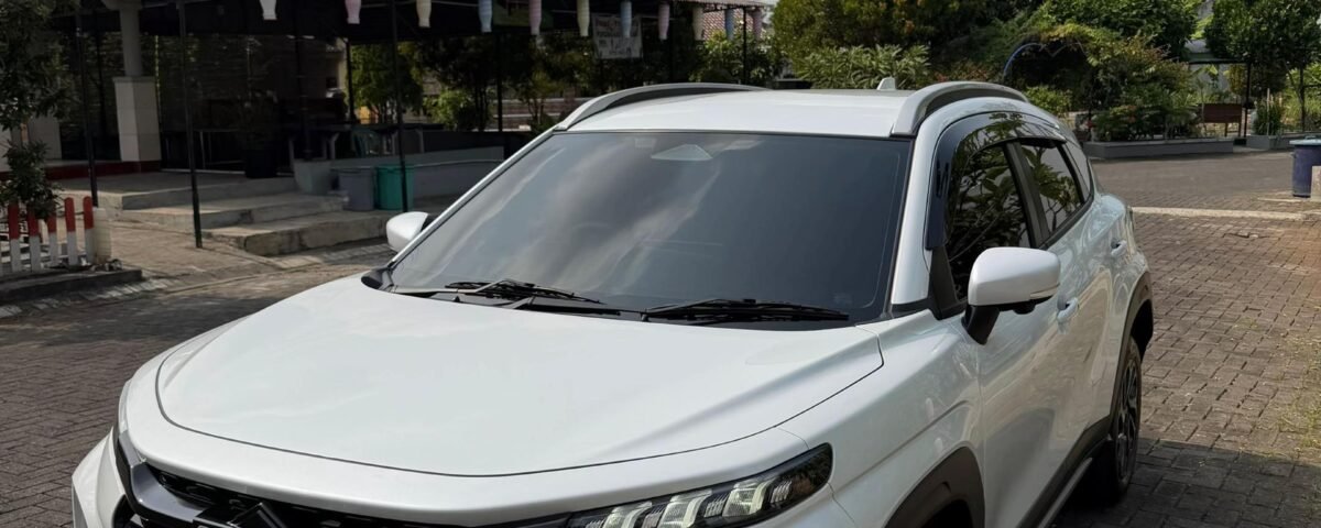 snaptik-app-7664784037243915541-slide-2 - Dyantransport White Suzuki SUV parked on a brick driveway in front of a shaded outdoor area with plants and a curved roof structure under a partly cloudy sky