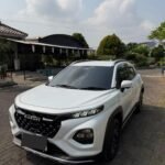 White Suzuki SUV parked on a brick driveway in front of a shaded outdoor area with plants and a curved roof structure under a partly cloudy sky