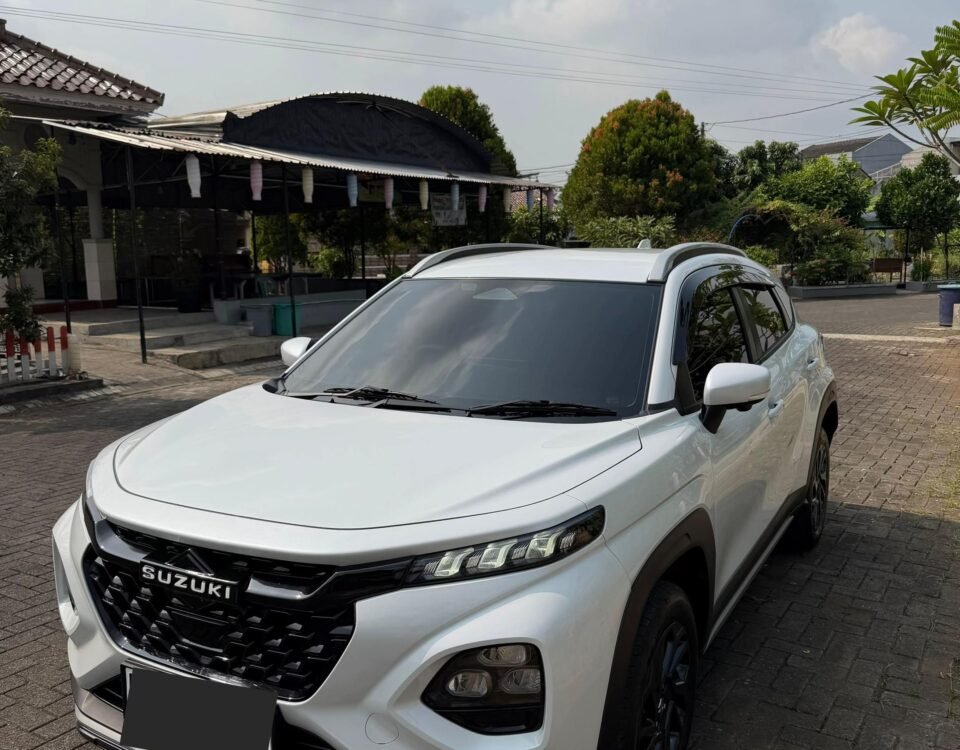 snaptik-app-7664784037243915541-slide-2 - Dyantransport White Suzuki SUV parked on a brick driveway in front of a shaded outdoor area with plants and a curved roof structure under a partly cloudy sky