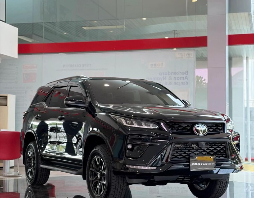 Black Toyota Fortuner on showroom floor, glossy finish, front grille visible, reflections on polished tiles behind glass walls