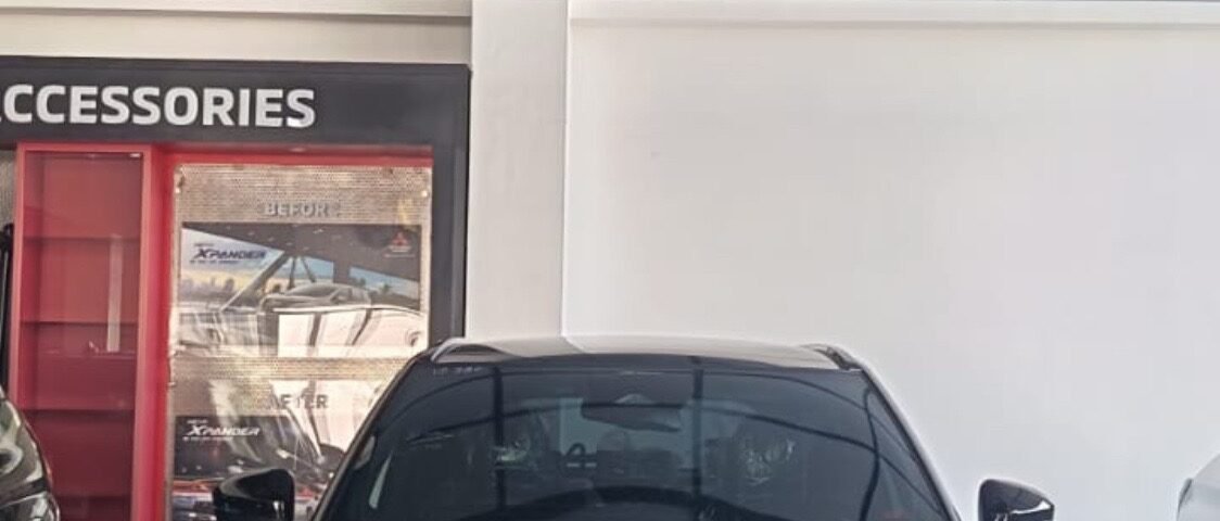 snaptik-app-7675198232921705749-slide-1 - Dyantransport Front view of a white Mitsubishi XFORCE SUV on a showroom floor, with a red garland overhead and accessories display to the left.