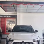 Front view of a white Mitsubishi XFORCE SUV on a showroom floor, with a red garland overhead and accessories display to the left.