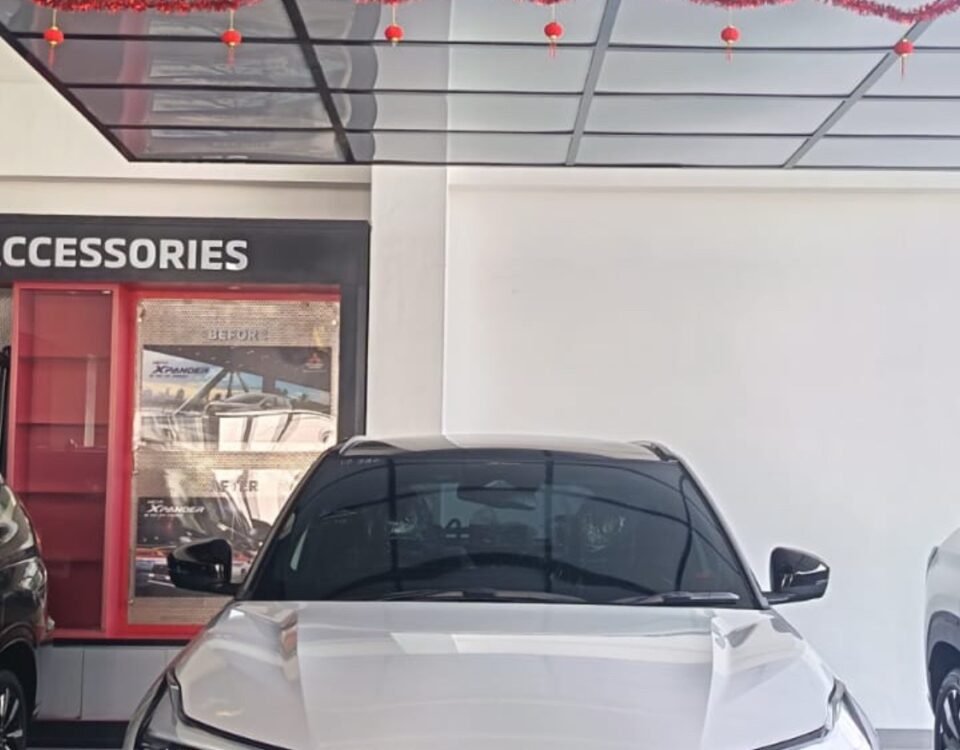 snaptik-app-7675198232921705749-slide-1 - Dyantransport Front view of a white Mitsubishi XFORCE SUV on a showroom floor, with a red garland overhead and accessories display to the left.