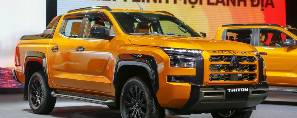 Orange Mitsubishi Triton pickup on display at a car show, with a second orange vehicle in the background and a colorful banner behind it.