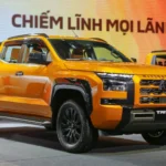 snaptik-app-7675633763791162632-slide-3 - Dyantransport Orange Mitsubishi Triton pickup on display at a car show, with a second orange vehicle in the background and a colorful banner behind it.