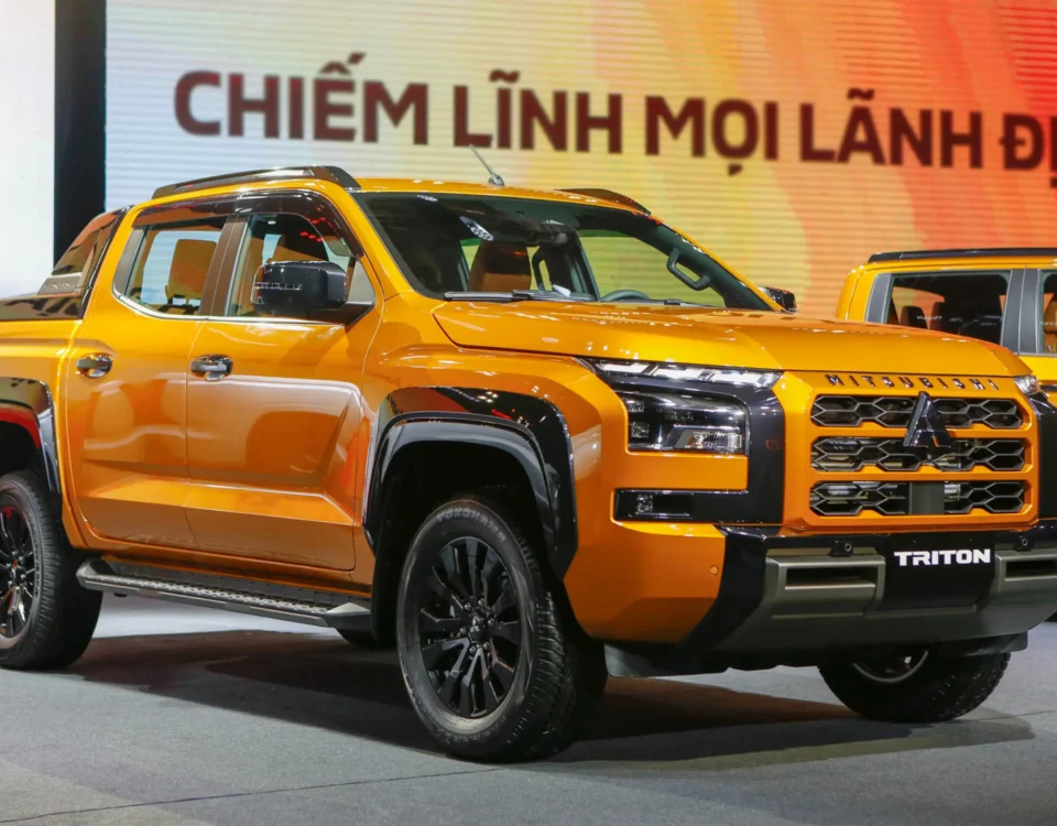 Orange Mitsubishi Triton pickup on display at a car show, with a second orange vehicle in the background and a colorful banner behind it.
